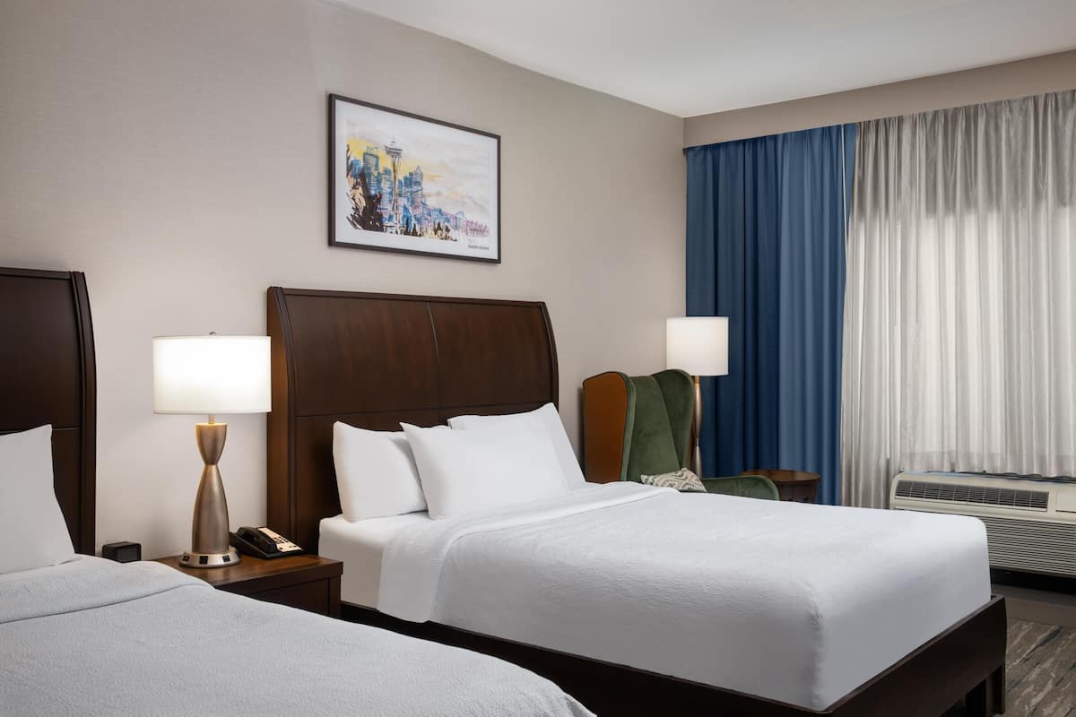 A hotel room with two beds, a nightstand with a lamp, a telephone, a painting on the wall, a chair, and a window with curtains.