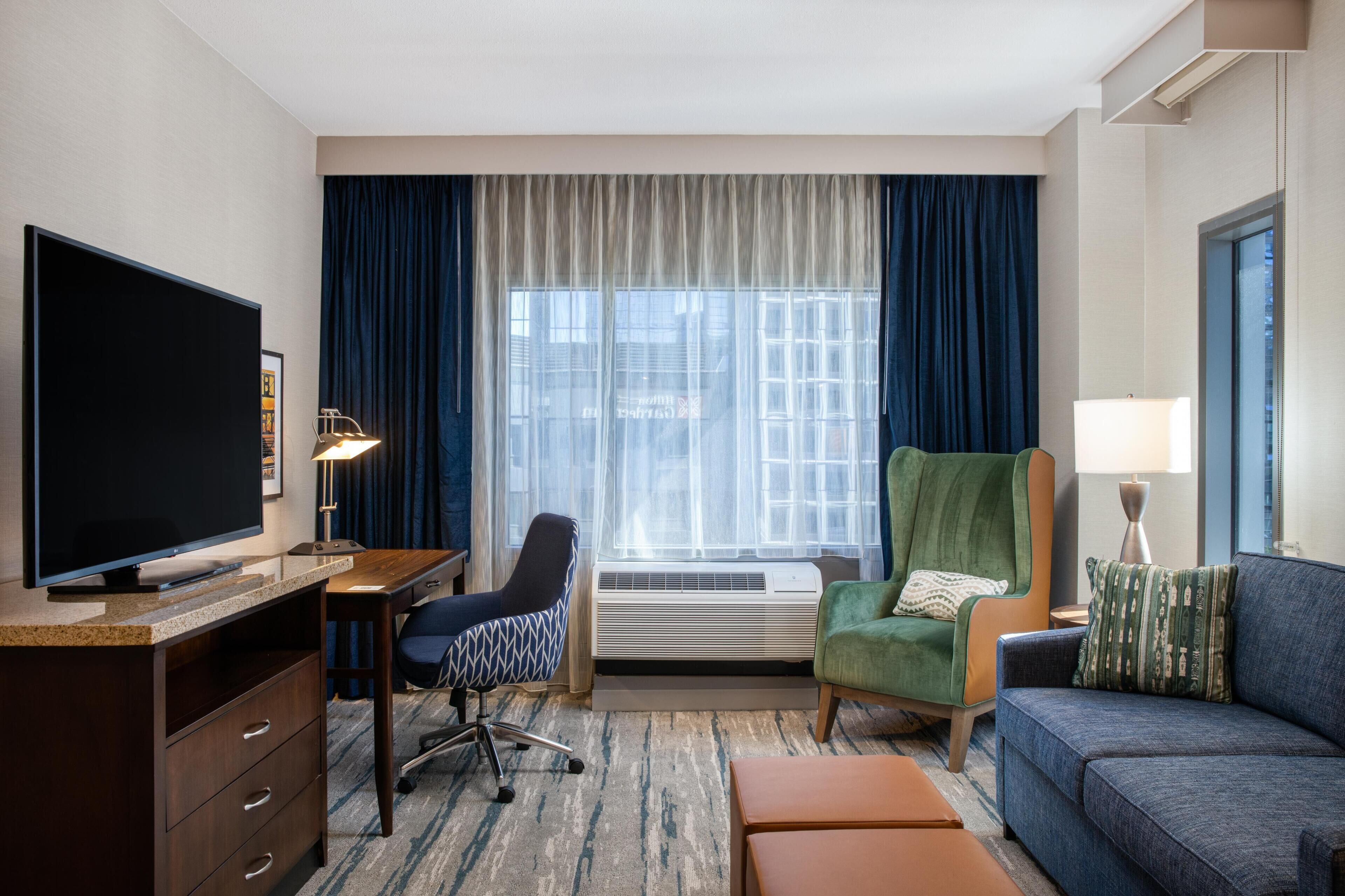 Habitación de hotel con televisión de pantalla plana, un escritorio, un sillón verde, un sofá azul y un ventanal con cortinas.