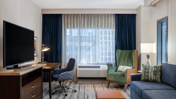 Habitación de hotel con televisión de pantalla plana, un escritorio, un sillón verde, un sofá azul y un ventanal con cortinas.