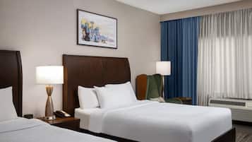 Habitación de hotel con dos camas, una mesita de noche con lámpara, teléfono, un cuadro en la pared, una silla y una ventana con cortinas.