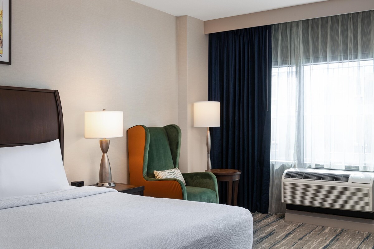 Ein Hotelzimmer mit einem Bett, zwei Nachttischlampen, einem Sessel, einem kleinen Tisch und einer Klimaanlage.