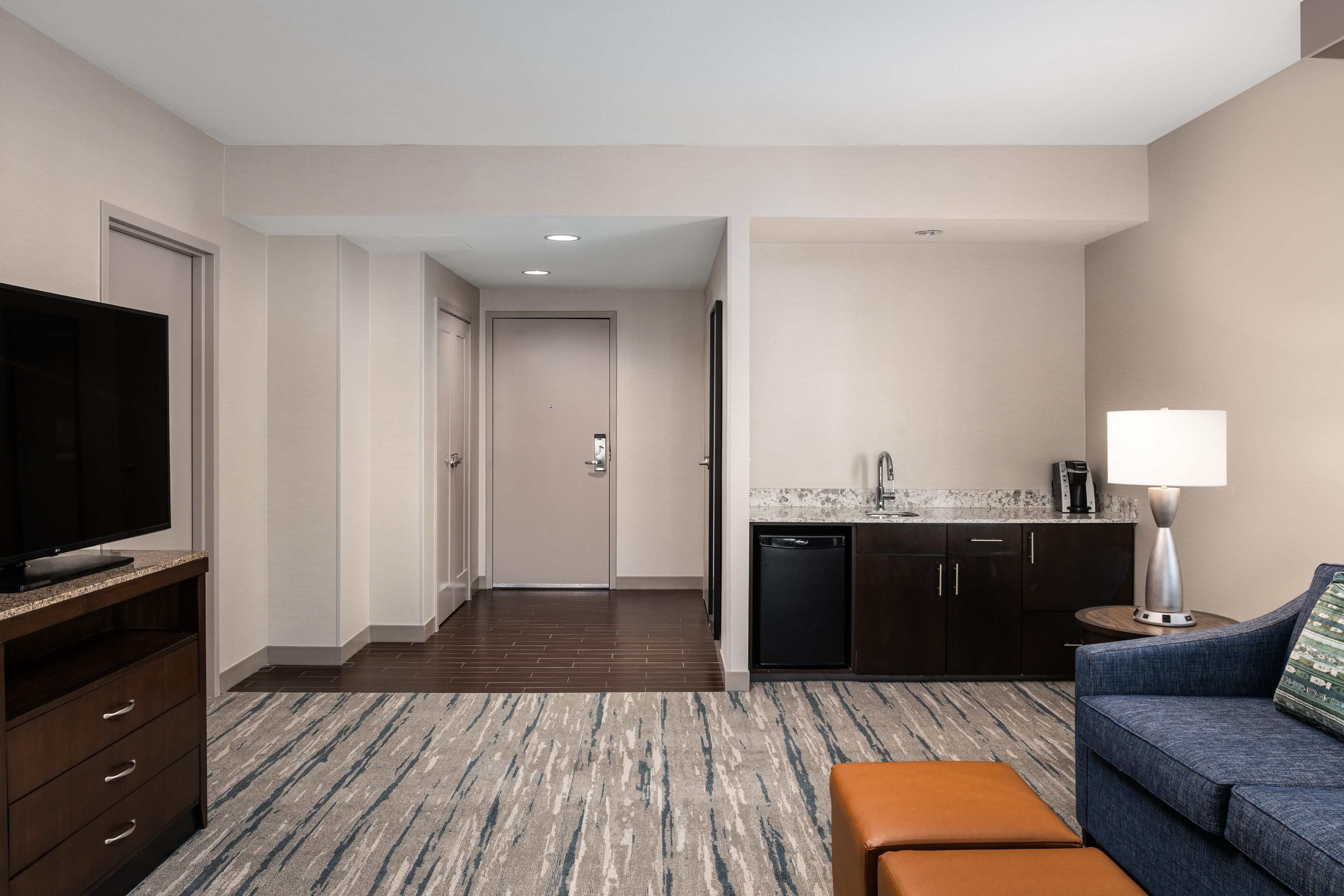 A hotel room with a television, sofa, ottoman, kitchen area, and a hallway leading to another room.