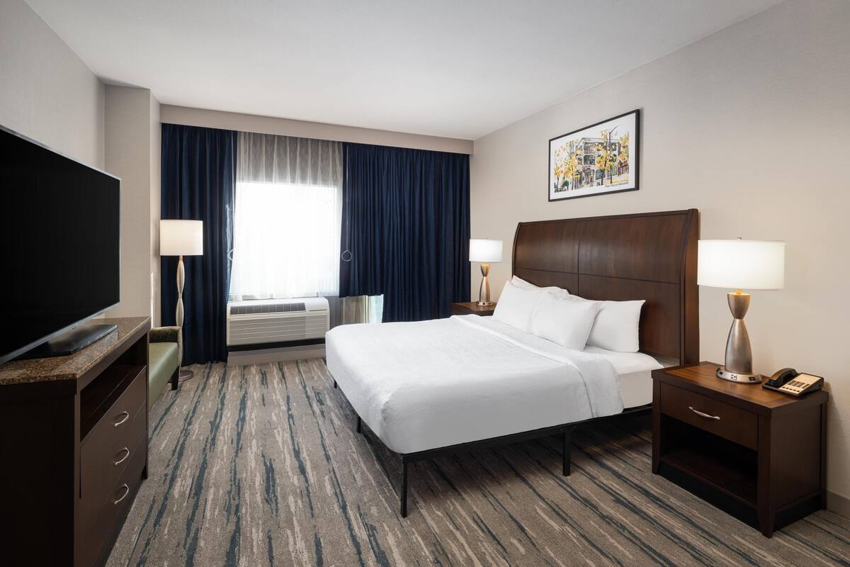 A hotel room with a bed, bedside tables, a TV, a window with curtains, and a wall-mounted artwork.