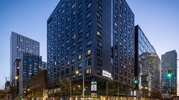 Un hotel de varios pisos con un llamativo letrero de Hilton Garden Inn, rodeado de edificios altos y una calle concurrida con semáforos.
