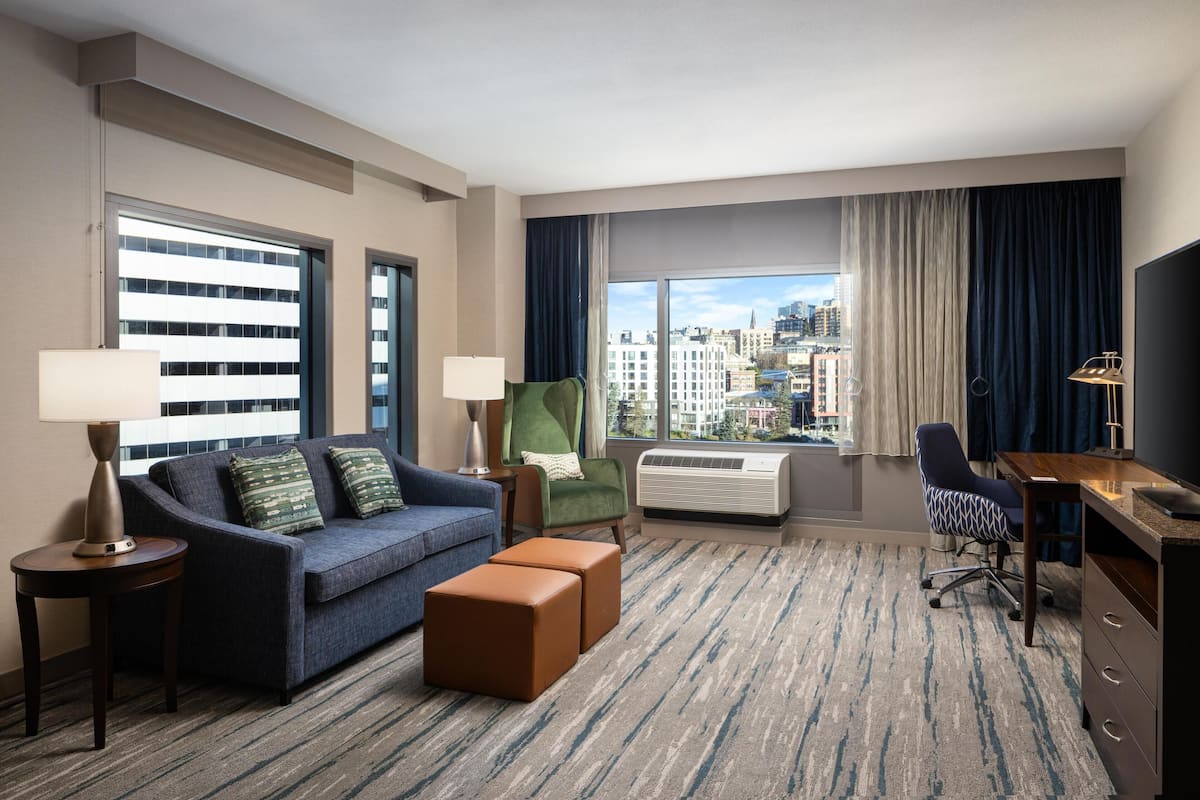 A hotel room with a sofa, armchair, desk, and large windows with a city view.