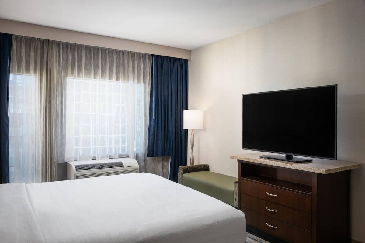A hotel room with a bed, a television on a stand, a desk with a lamp, and an air conditioning unit.