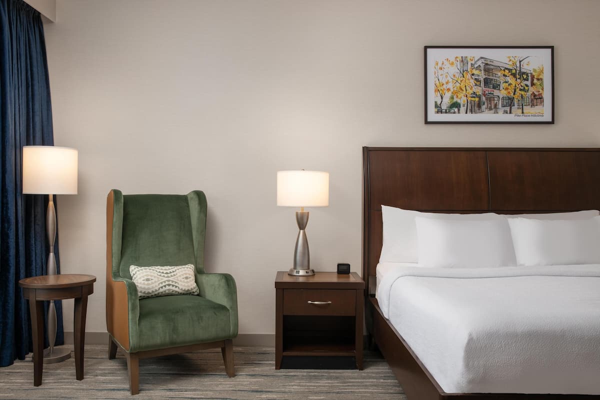A hotel room with a bed, a green armchair, a nightstand with a lamp, a framed picture on the wall, and a small table.