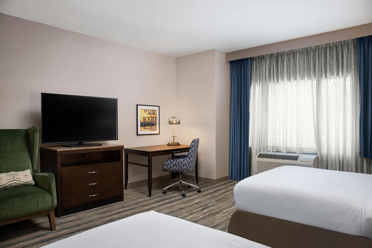 A hotel room with a flat-screen TV, a desk with a lamp, a patterned chair, two beds, and a large window with curtains.
