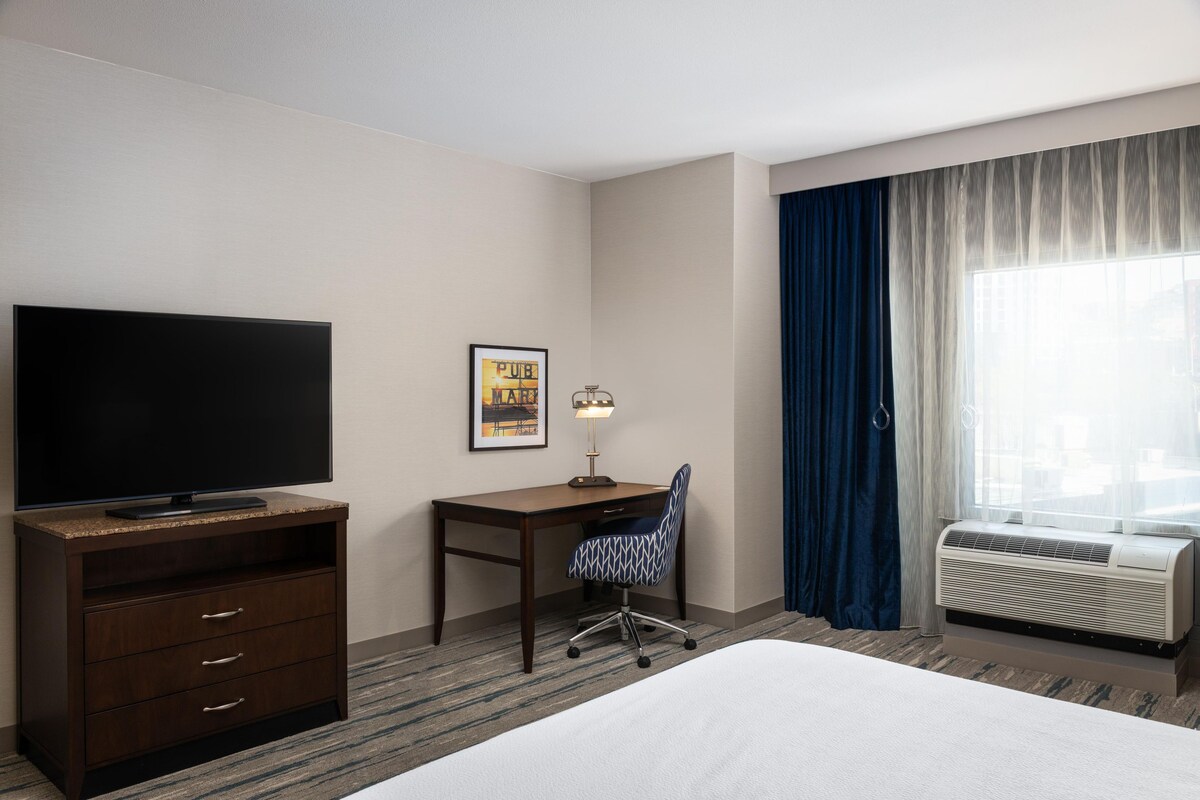 Ein Hotelzimmer mit Bett, Schreibtisch mit Stuhl, Fernseher, einem gerahmten Bild, einer Klimaanlage und einem Fenster mit Vorhängen.