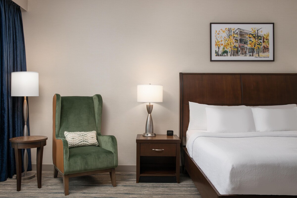 Ein Hotelzimmer mit einem Bett, einem grünen Sessel, einem Nachttisch mit Lampe, einem kleinen Tisch und einem gerahmten Bild an der Wand.