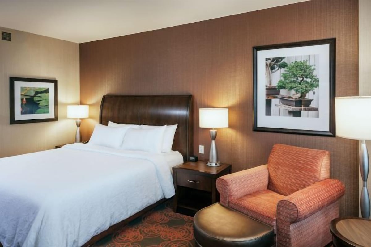 A hotel room with a large bed, a nightstand, a chair, and two framed pictures on the wall.