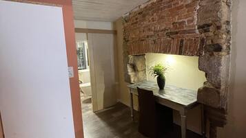 Een smalle gang met een bakstenen muur en een houten deur. Tegen de muur staat een klein tafeltje met een bloempot.