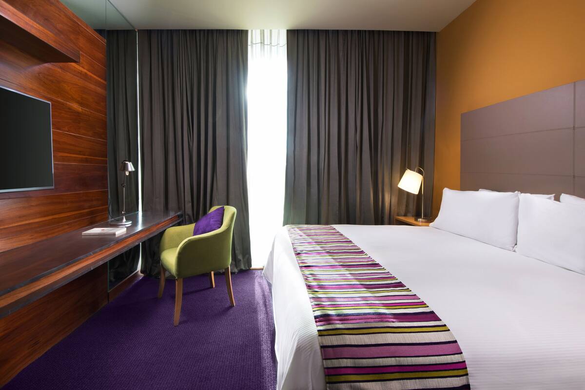 Una habitación de hotel con una cama, un escritorio con silla, un televisor y una ventana con cortinas.