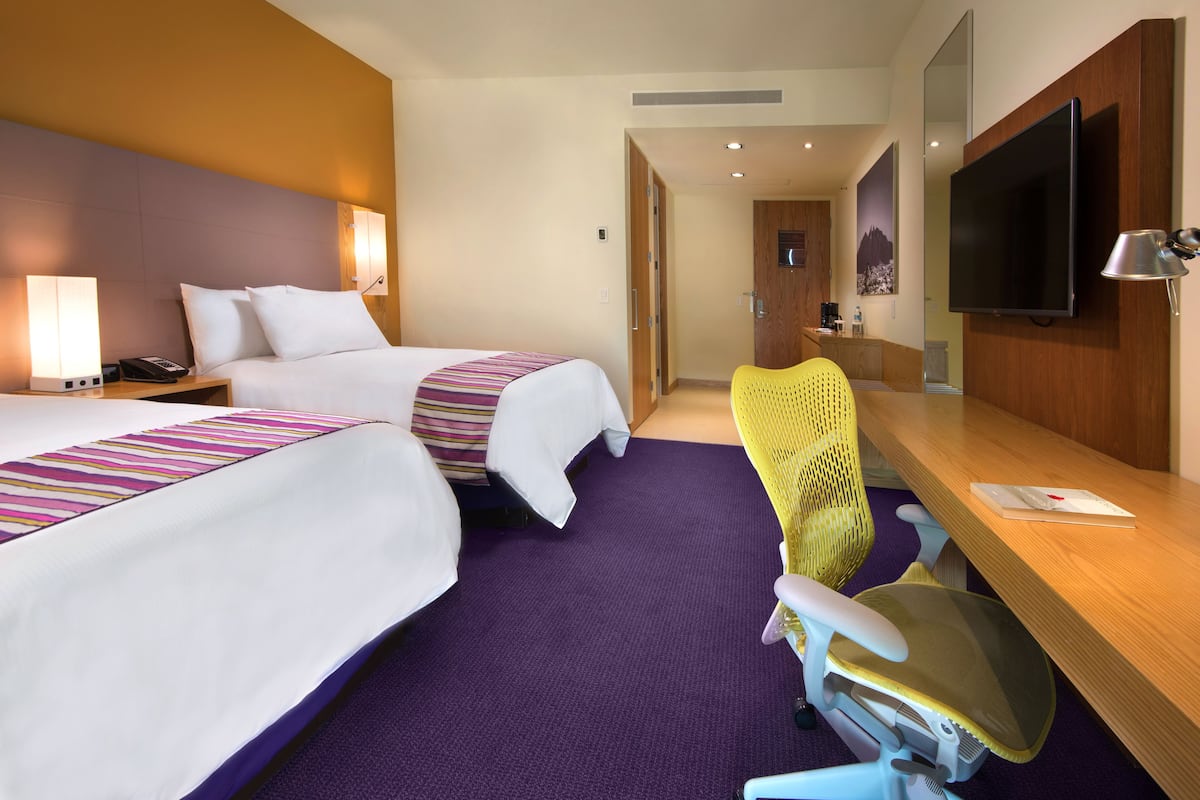 Habitación de hotel con dos camas, un escritorio con silla, televisor y baño visible a través de una puerta abierta.