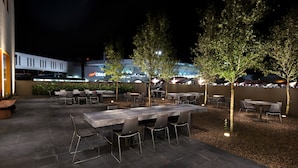 Un patio al aire libre con mesas y sillas, árboles y un edificio con señalización iluminada al fondo.