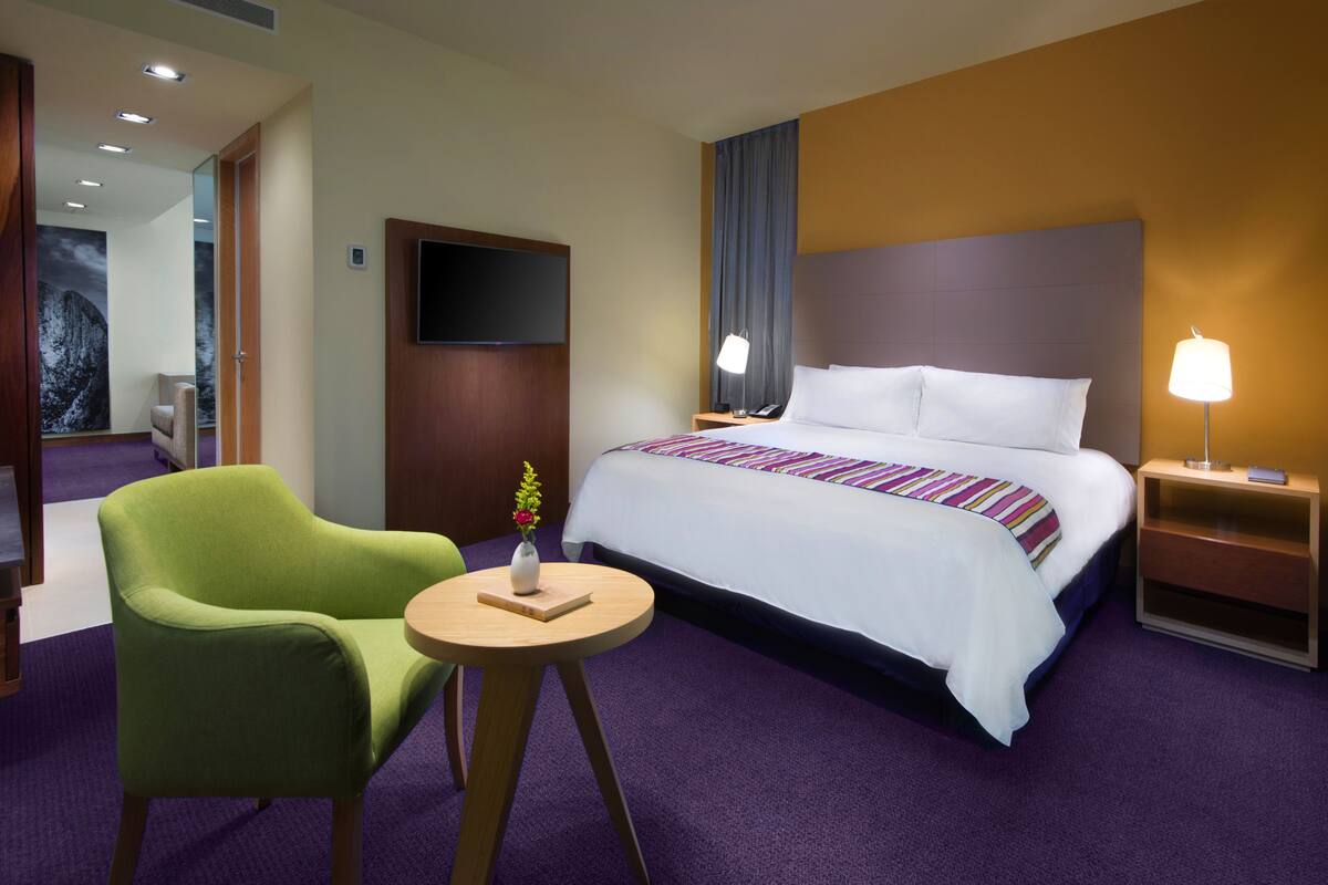 Una habitación de hotel con una cama grande, un sillón verde, una mesita redonda con un florero y un televisor montado en la pared.