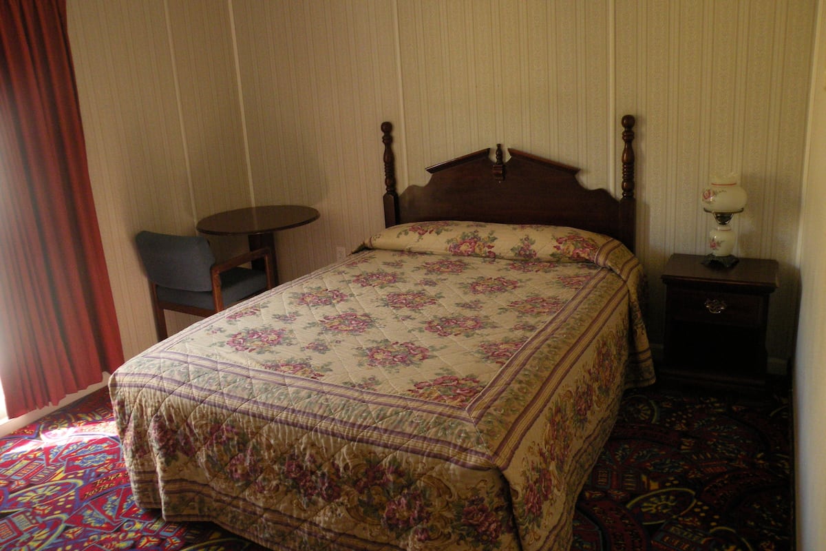 A bedroom with a wooden bed, a small round table, a chair, and a nightstand with a lamp.