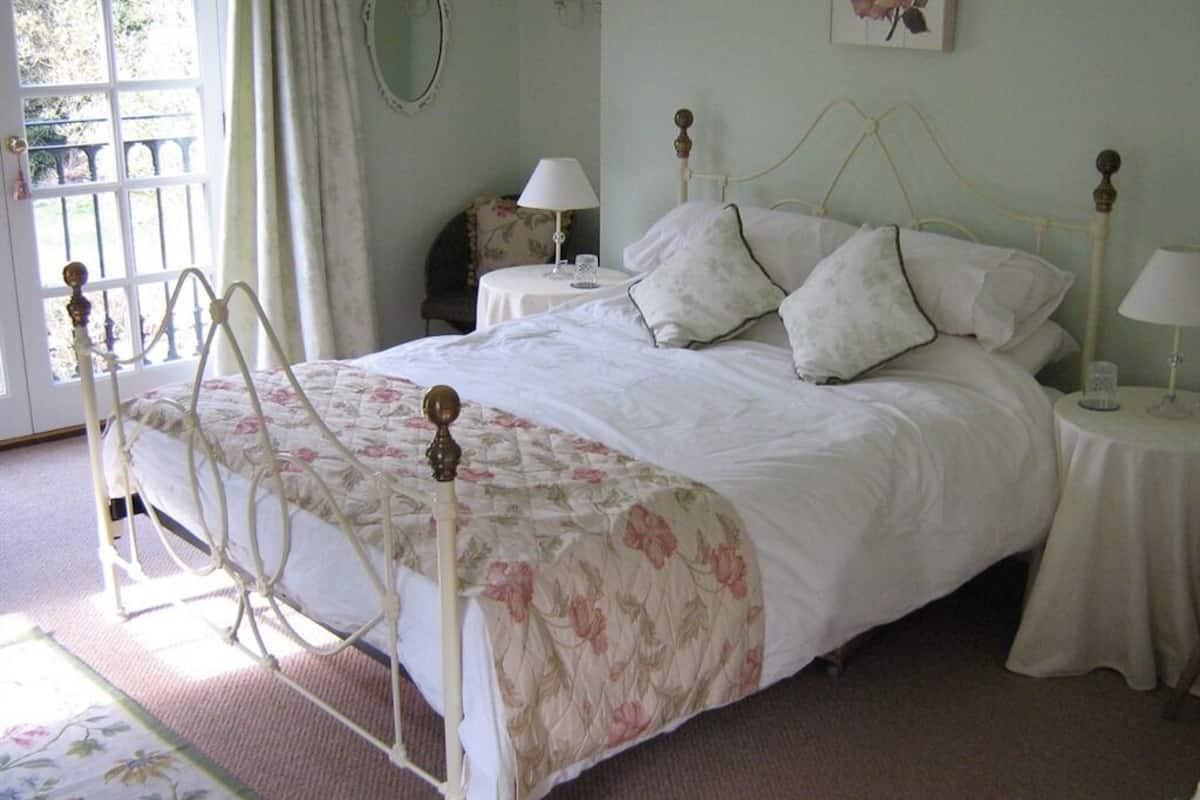 A bedroom with a four-poster bed, two bedside tables with lamps, a floral-patterned bedspread, and a view of the outdoors through a window with curtains.