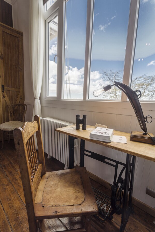 Una habitación rústica con una silla de madera, un escritorio con una lámpara antigua y vistas del cielo a través de una ventana.
