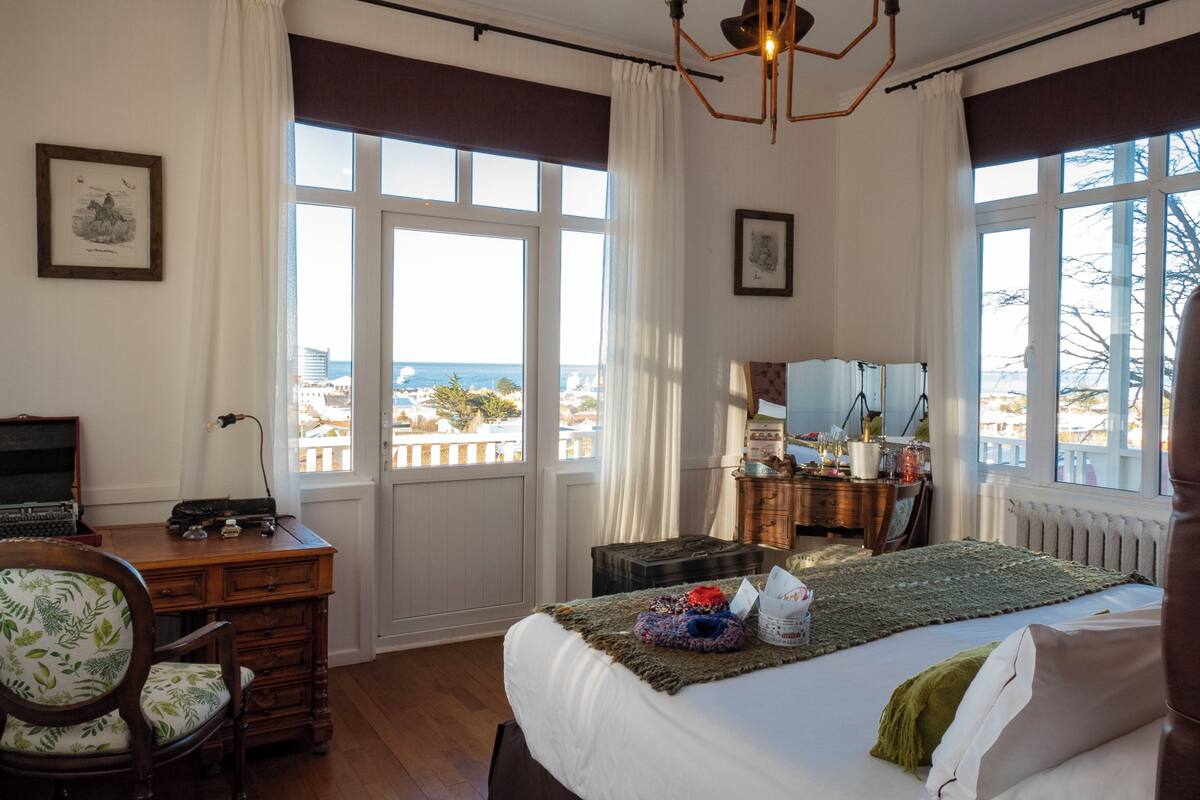 A bedroom with a bed, a desk, a chair, a dresser, and a view of the sea.