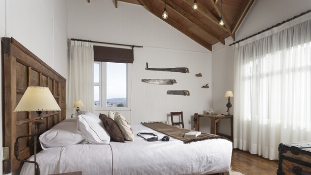 Un dormitorio con cabecera de madera, cama con ropa de cama blanca, mesita de noche de madera con lámpara, escritorio con silla y un ventanal con cortinas.