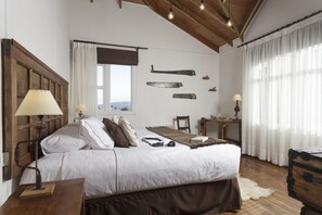 Un dormitorio con cabecera de madera, cama con ropa de cama blanca, mesita de noche de madera con lámpara, escritorio con silla y un ventanal con cortinas.