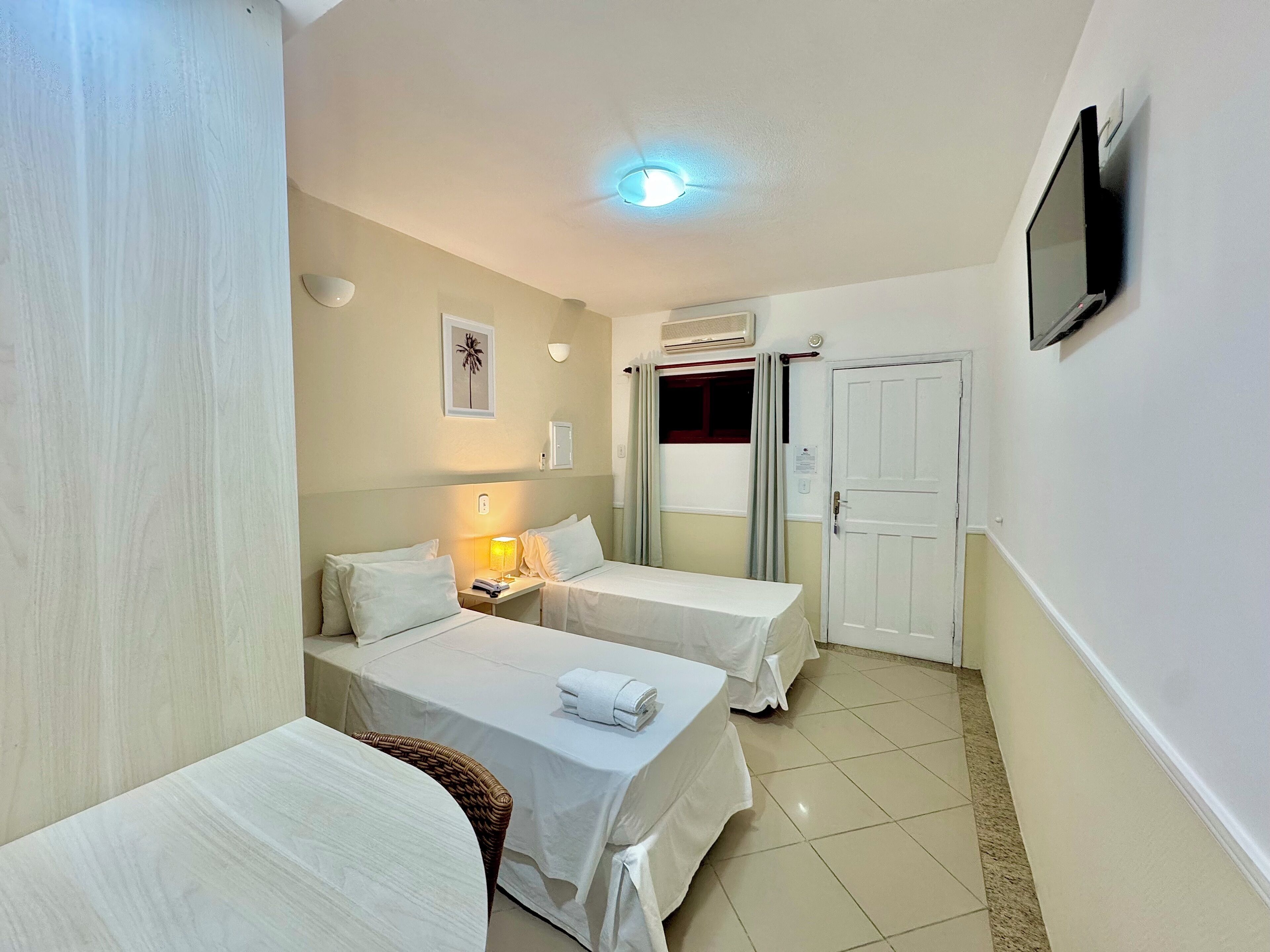 A hotel room with two single beds, a bedside table with a lamp, a TV mounted on the wall, and a window with curtains.