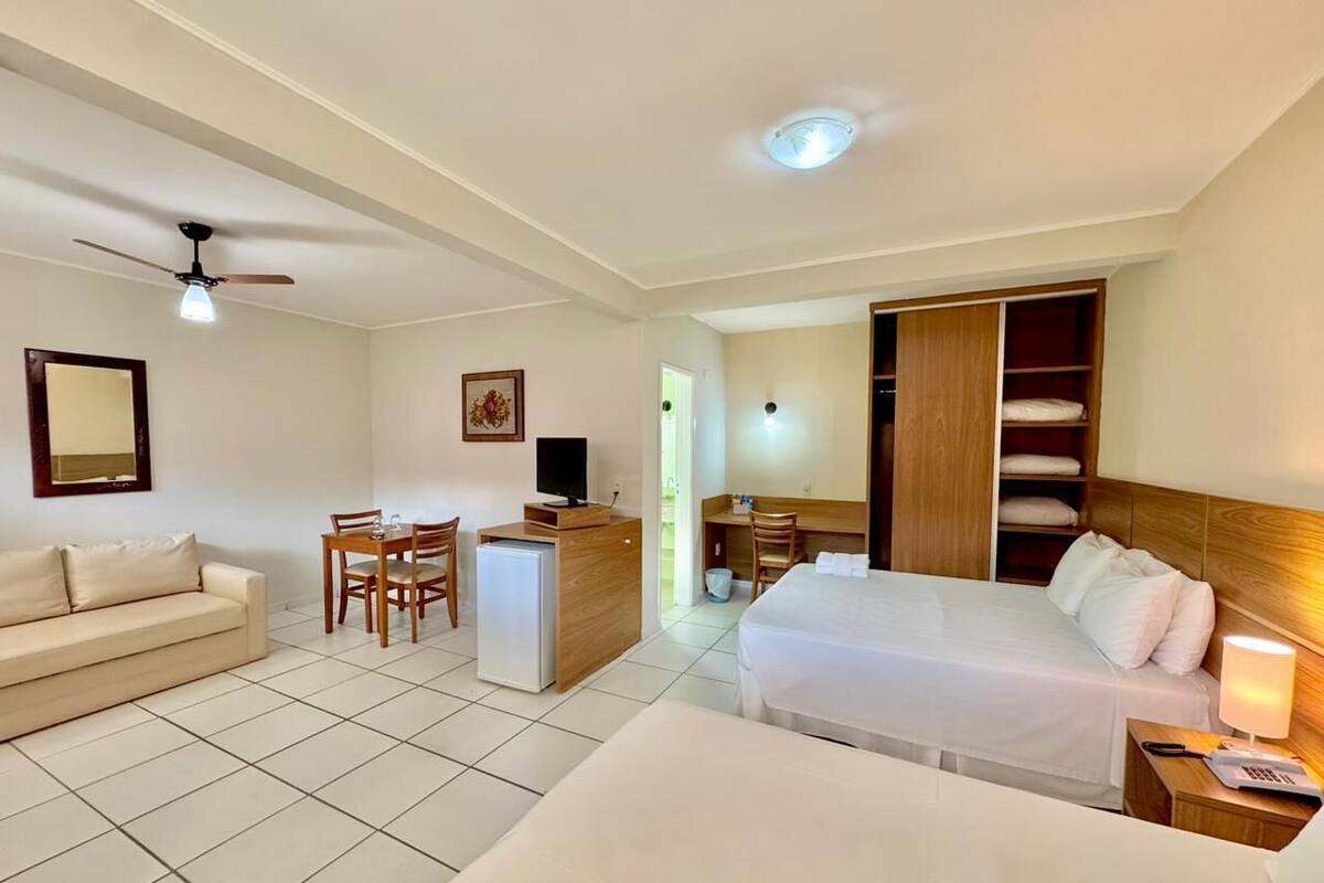 A hotel room with a bed, a desk, a chair, a small fridge, a TV, a ceiling fan, and a wardrobe.
