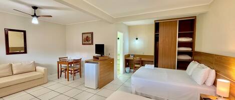 A hotel room with a bed, a desk, a chair, a small fridge, a TV, a ceiling fan, and a wardrobe.