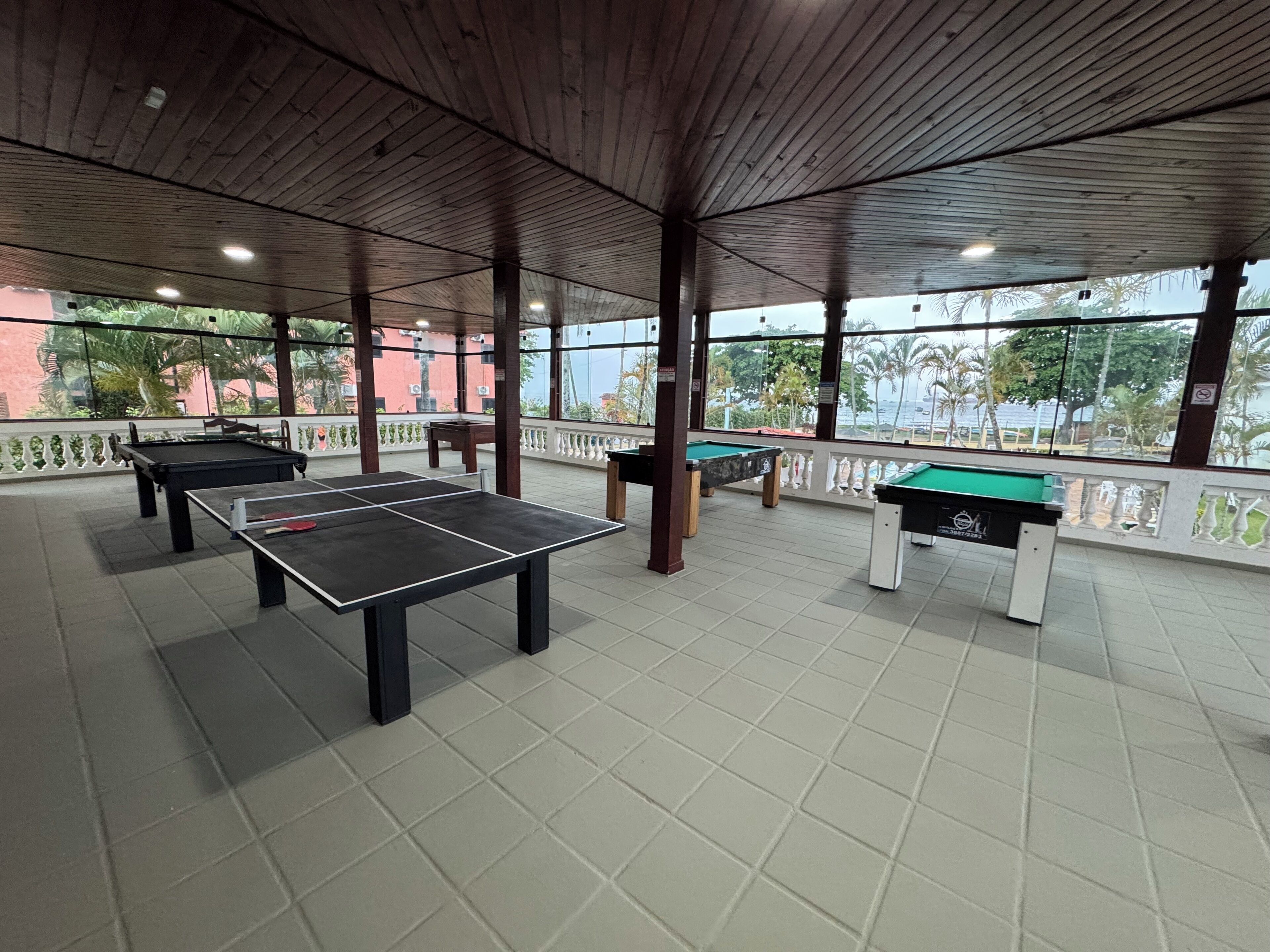 An indoor recreational area with multiple foosball tables and a pool table, featuring a wooden ceiling and large windows overlooking a landscaped area.