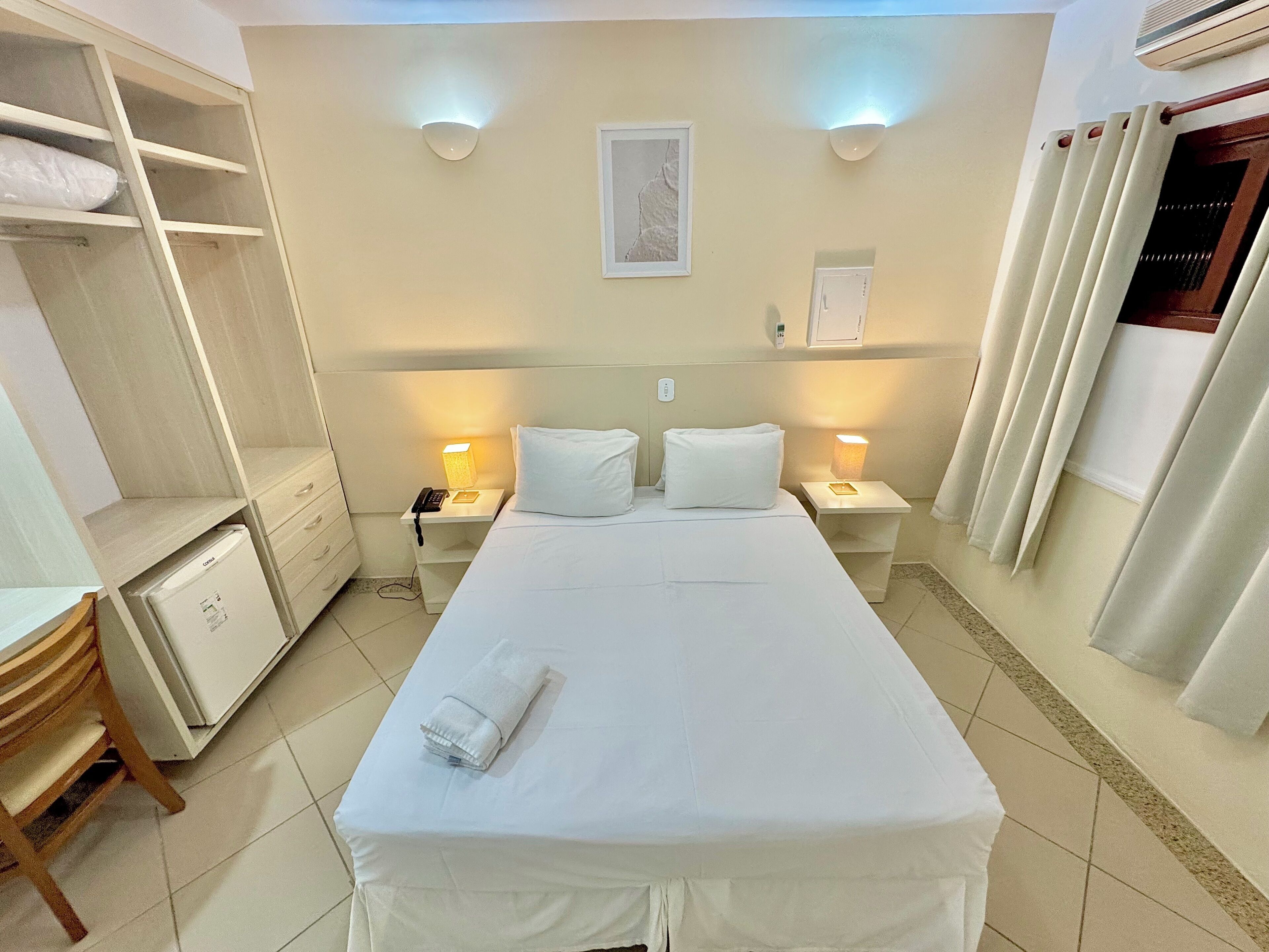 Una habitación de hotel con una cama, dos mesitas de noche con lámparas, un armario, un escritorio y un refrigerador pequeño.
