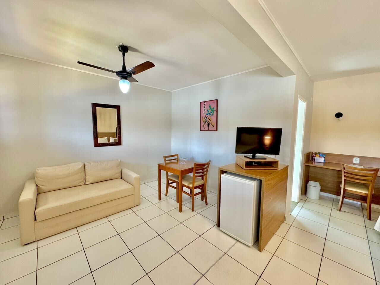 Um espaço compacto com sofá, mesa de jantar com cadeiras, pequena geladeira, televisão e ventilador de teto.