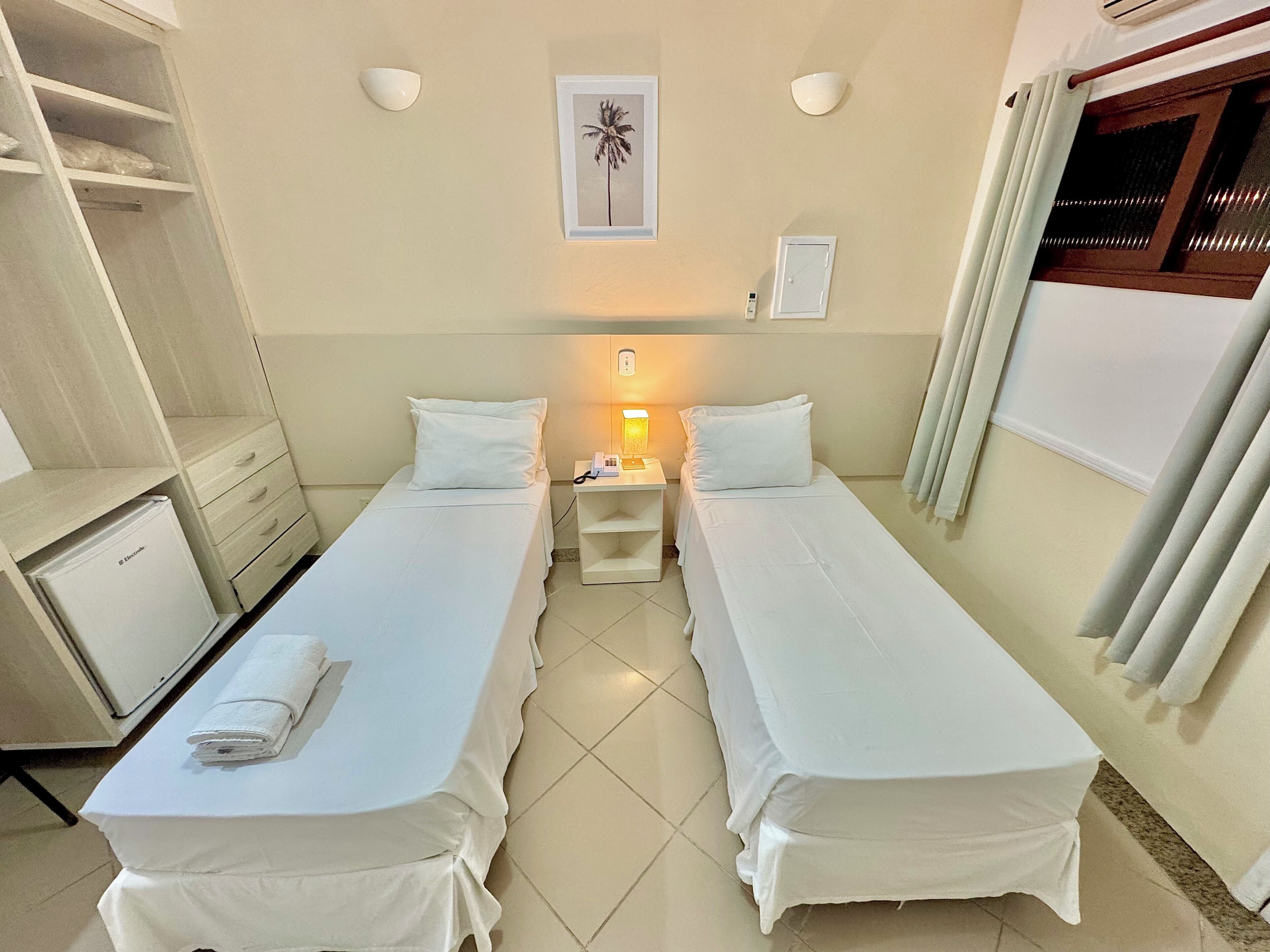 Une chambre d’hôtel avec deux lits simples, un bureau, une chaise, un petit réfrigérateur et un luminaire fixé au mur.