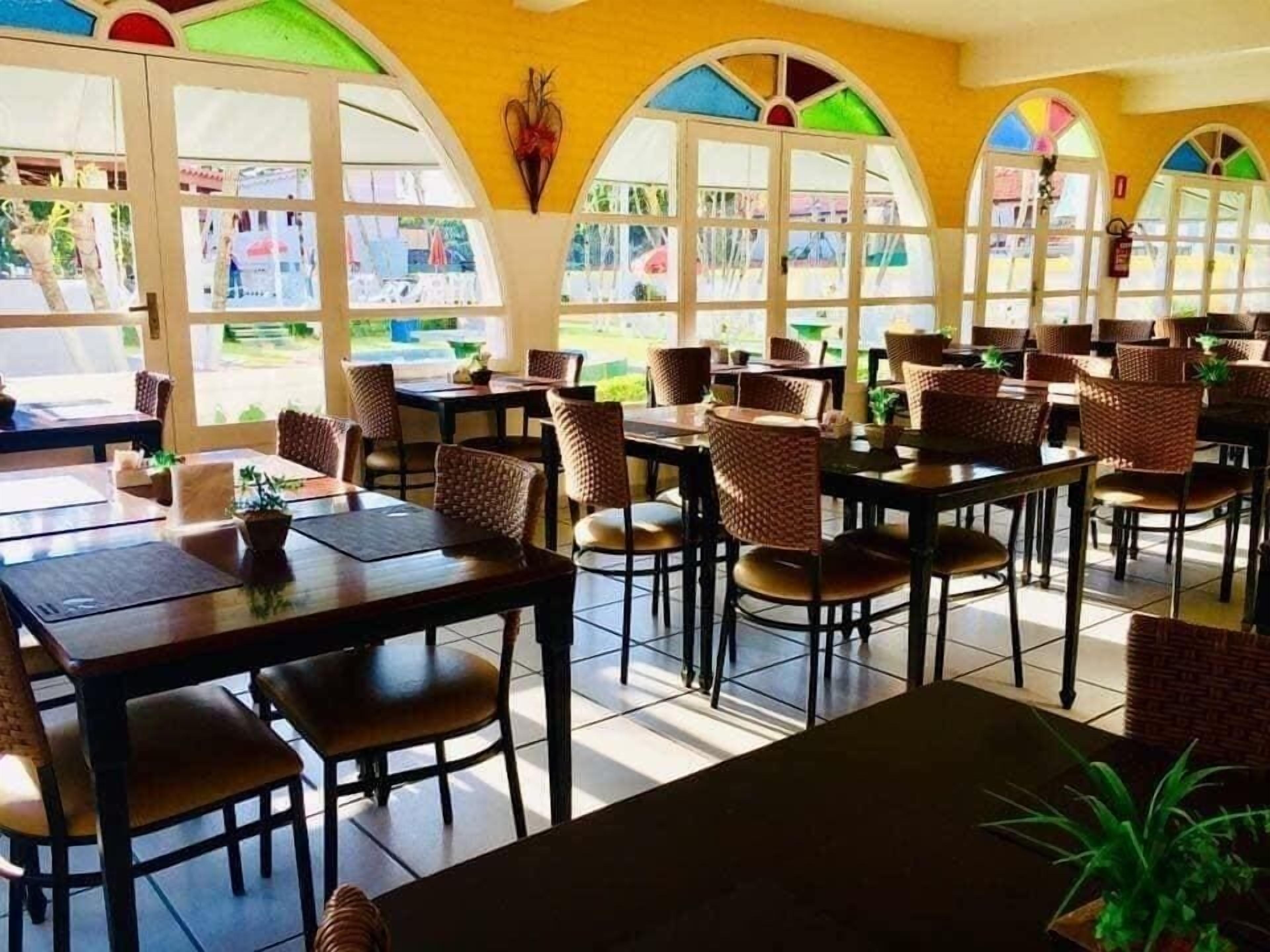 A restaurant with multiple tables and wicker chairs, large windows with colorful glass panes, and a tiled floor.