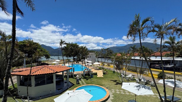 A pool area with a small building, palm trees, and a view of mountains. - Hotel Recanto dos Pássaros (São Sebastião)