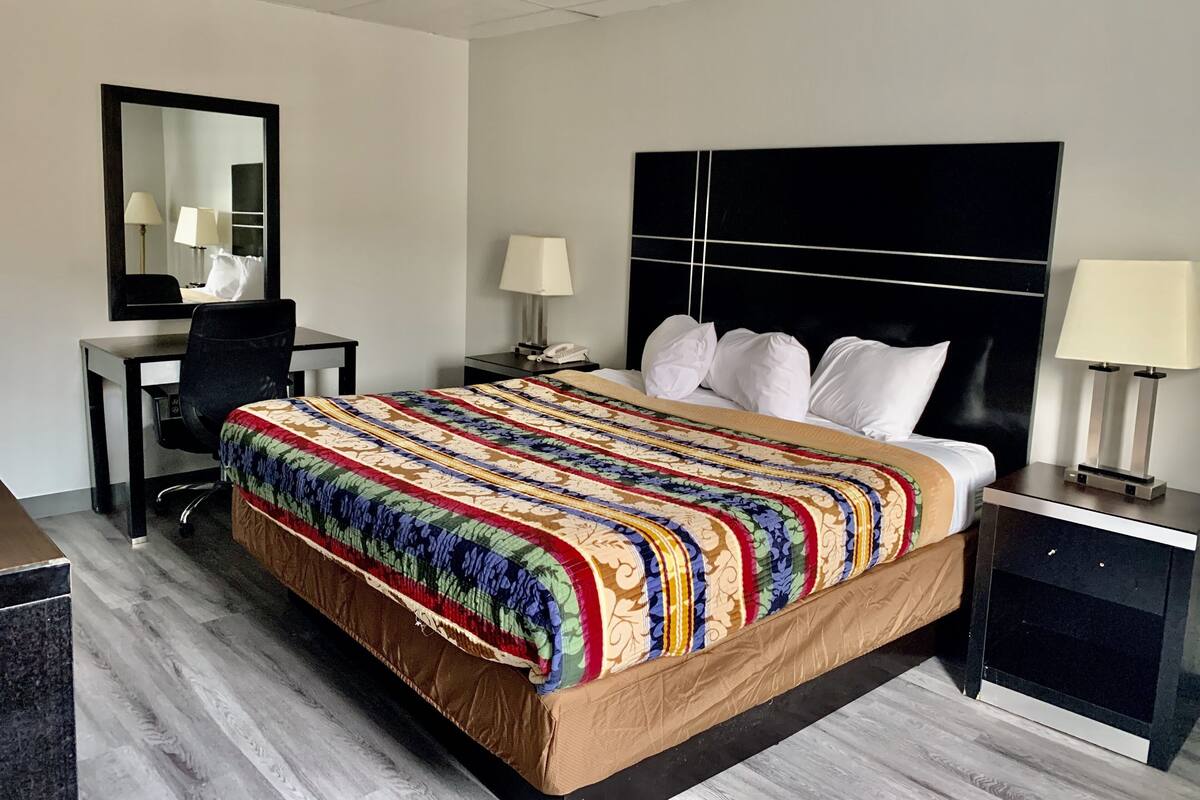 A hotel room with a bed, a desk, a chair, a nightstand, a lamp, and a mirror.