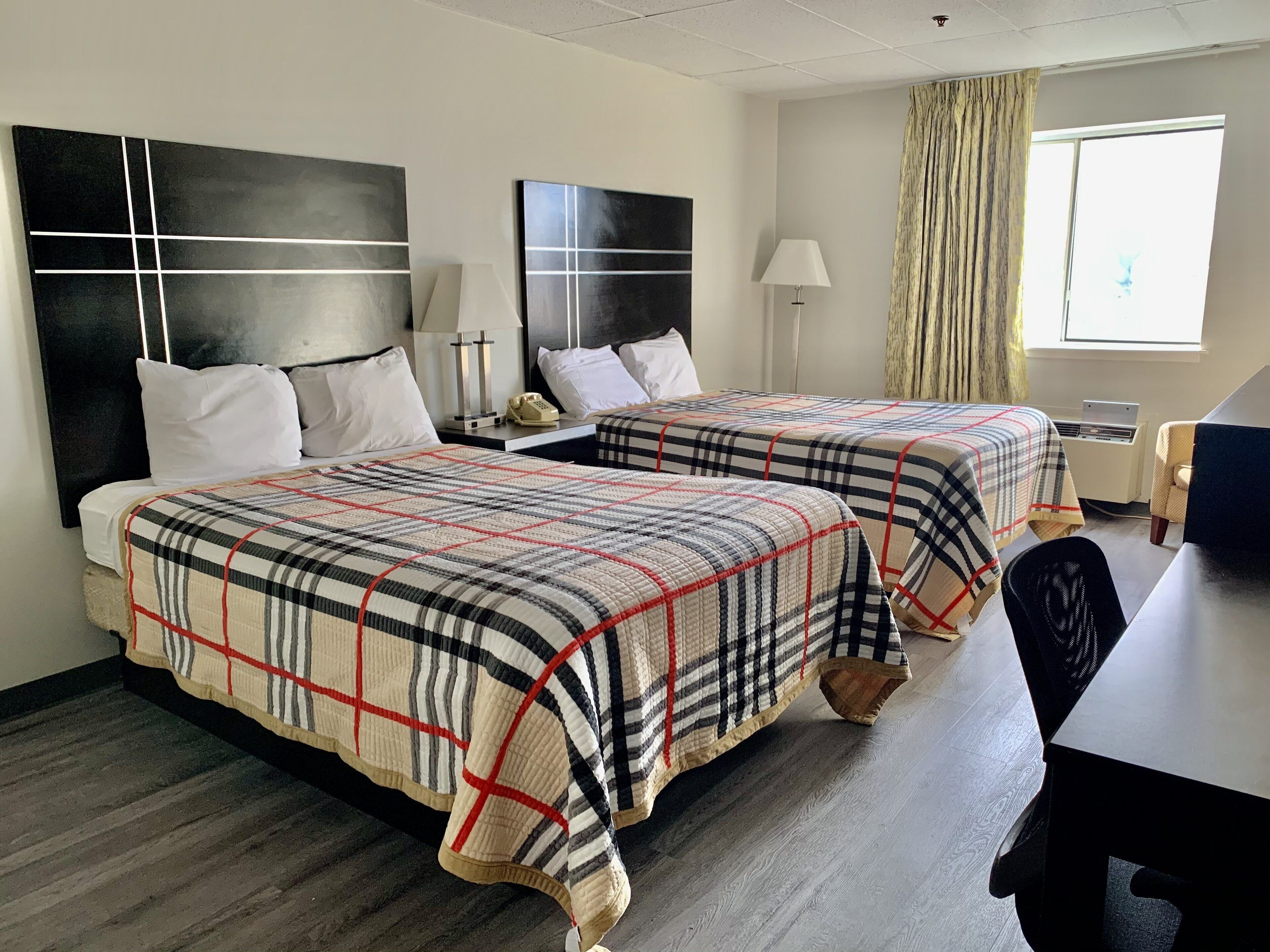 A hotel room with two beds, each with a plaid bedspread, a desk with a chair, a lamp, and a window with curtains.