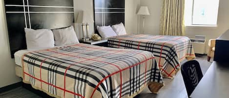 A hotel room with two beds, each with a plaid bedspread, a desk with a chair, a lamp, and a window with curtains.