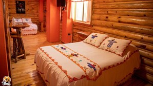 A cozy log cabin room with a large bed, a small table, and a window with curtains. - Hotel Taramuri (Bocoyna)