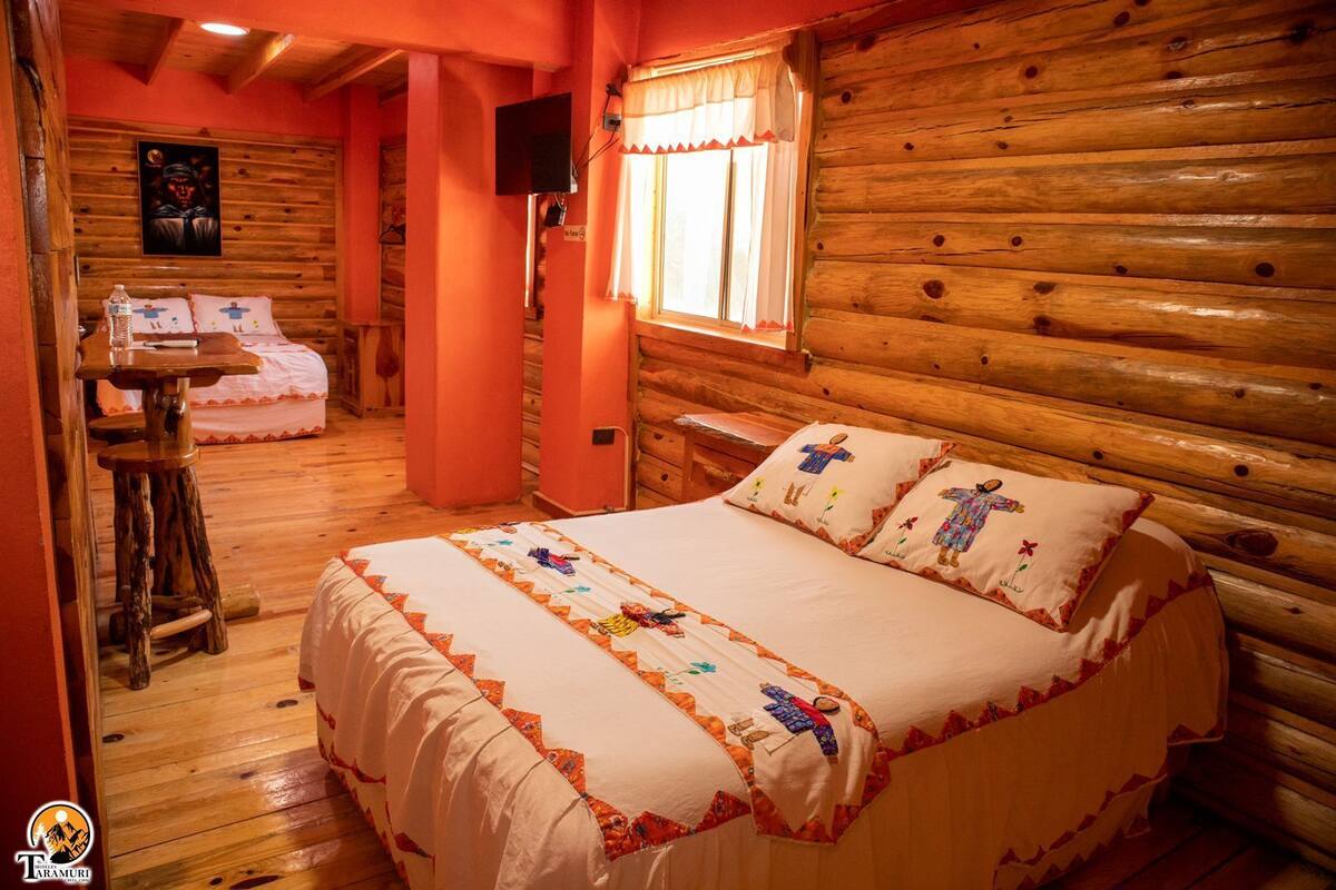 A cozy log cabin room with a large bed, a small table, and a window with curtains.