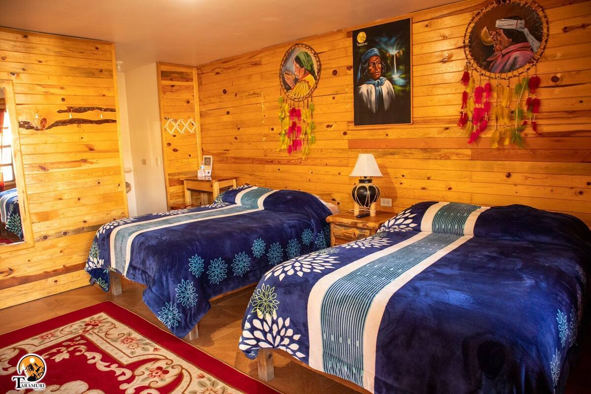 A wooden cabin room with two beds, a red carpet, and wall decorations including paintings and dreamcatchers.