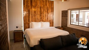 A bedroom with a wooden headboard, a large bed, a nightstand, a window with a view, and a logo in the corner. - Hotel Taramuri (Bocoyna)
