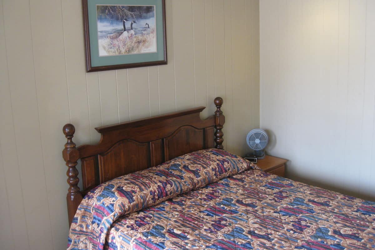 A bedroom with a wooden bed, a patterned bedspread, a framed painting of birds, and a standing fan.