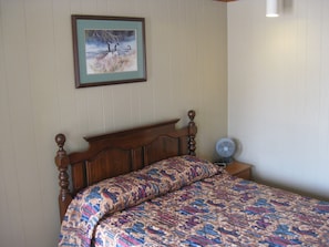 A bedroom with a wooden bed, a patterned bedspread, a framed painting of birds, and a standing fan. - Inn Town Motel (Waupun)