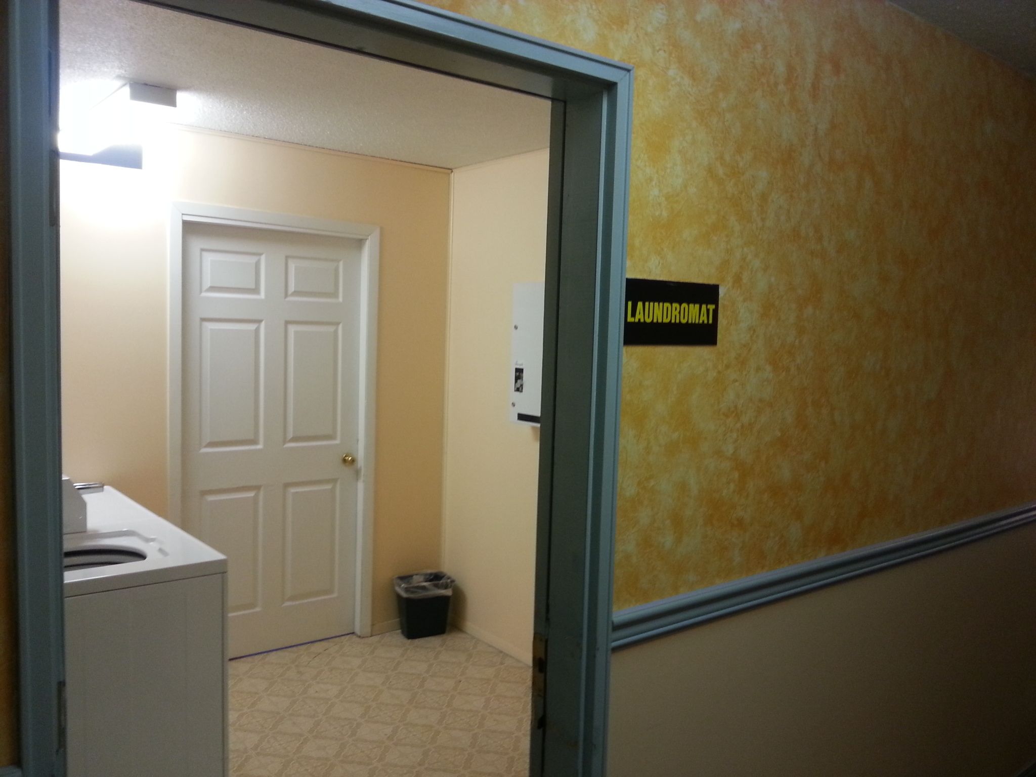 A hallway with a door leading to a room, a trash bin, and a sign that reads "LAUNDROMAT".