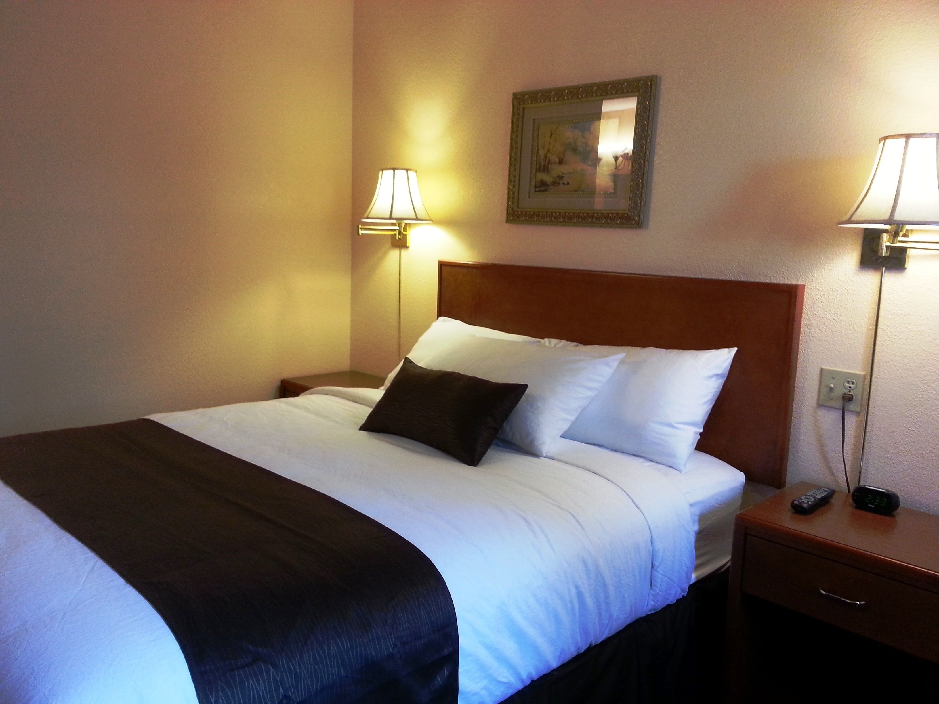 A neatly made bed with white and dark bedding, two bedside lamps, a nightstand with a remote control, and a framed picture on the wall.