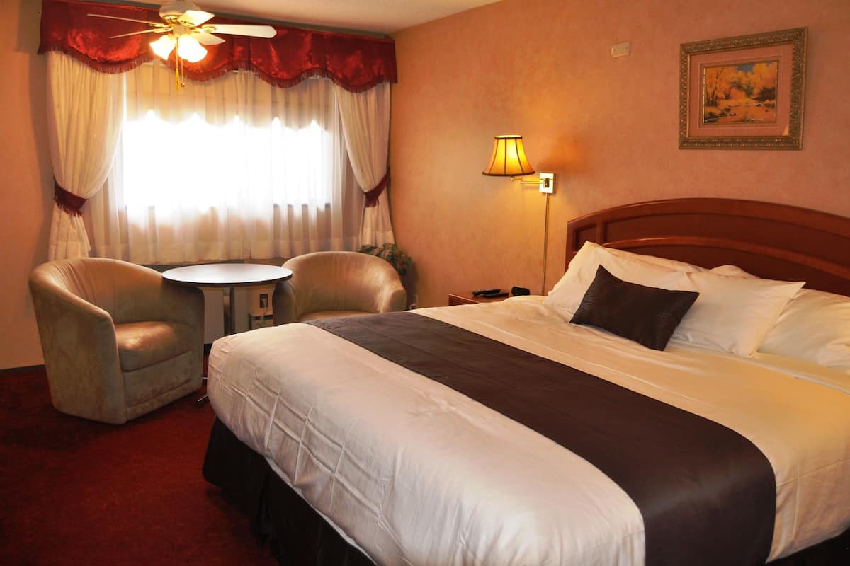 A hotel room with a large bed, two armchairs, a small round table, a ceiling fan, a lamp, and a painting on the wall.