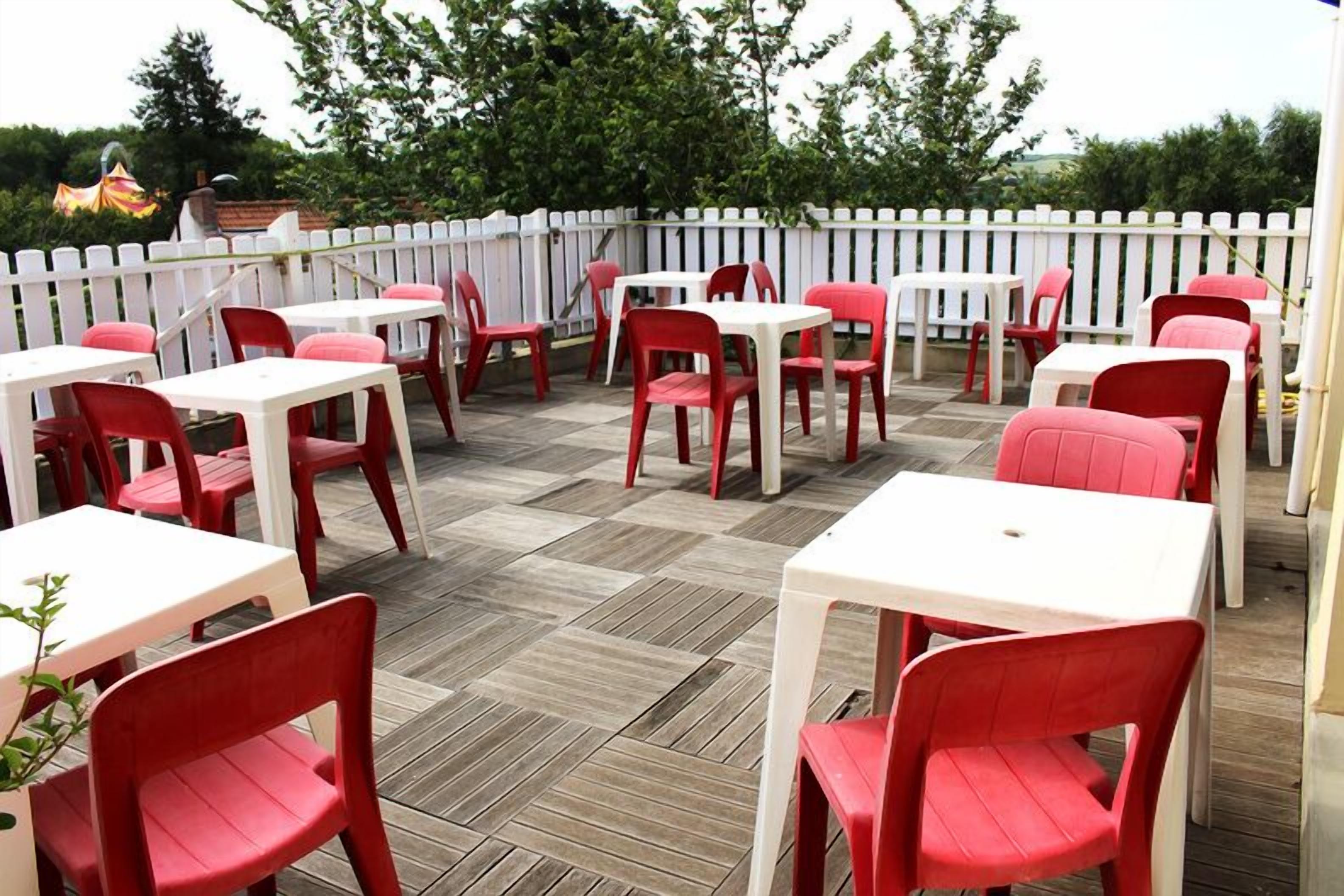 An outdoor patio with white tables and red chairs, surrounded by a white picket fence.