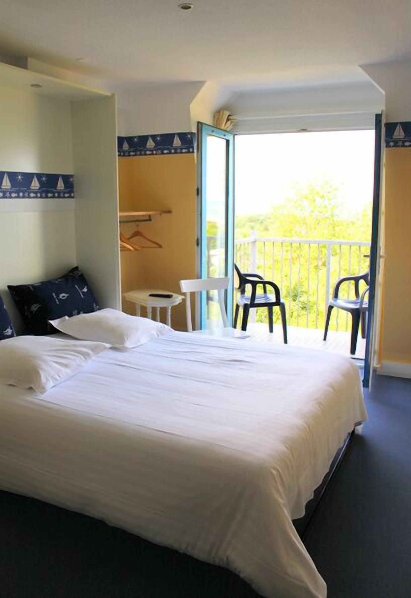 A hotel room with a large bed, a small table, two chairs, and a balcony with a railing.