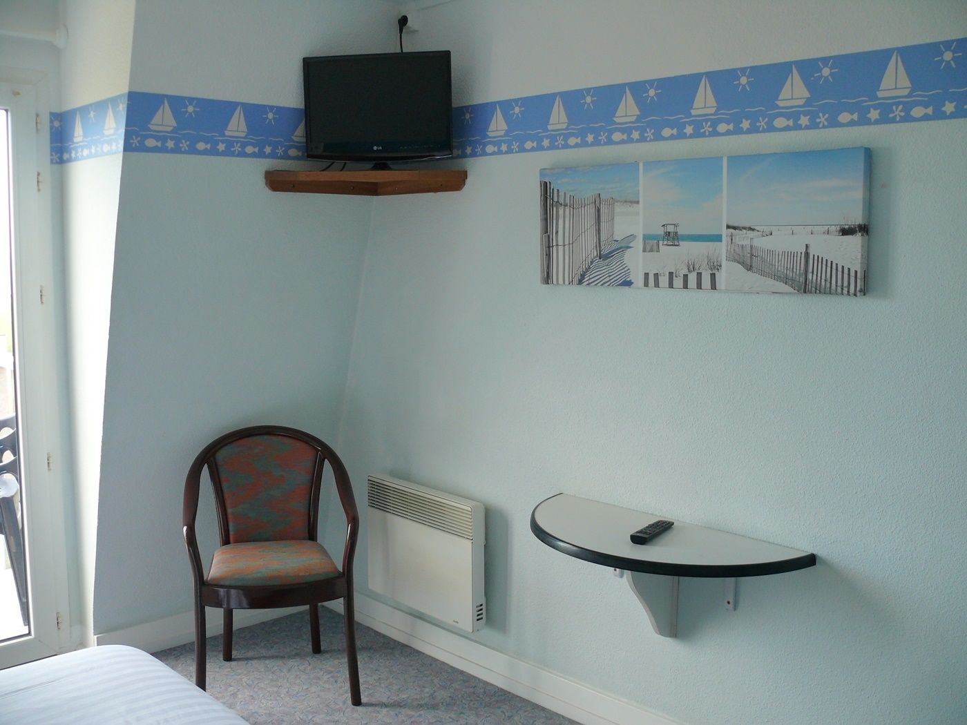 Habitación de hotel con una cama, una silla, una mesita, un televisor montado en un estante de madera y dos cuadros colgados en la pared.